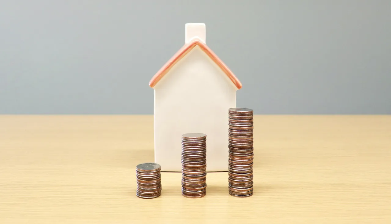 Miniatura de casa branca ao lado de pilhas de moedas sobre mesa de madeira