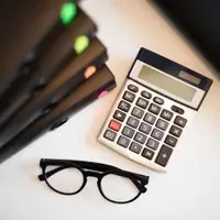 Calculadora de mesa ao lado de óculos de grau e de uma pilha de pastas de arquivo com marcadores coloridos, sobre superfície branca