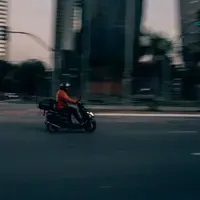Entregador de capacete e jaqueta laranja pilota uma moto com bolsa de entrega em uma avenida larga, com prédios e árvores desfocados pelo movimento, no fim da tarde