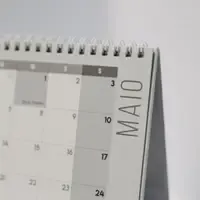 Calendário de mesa em espiral, aberto na página de maio em português, sobre uma parede clara desfocada