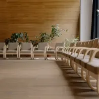 Salão vazio com fileiras de cadeiras de madeira clara e assento bege voltadas para uma parede revestida de ripas de madeira, com vasos de plantas entre as cadeiras da frente