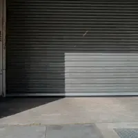 Porta de aço cinza de um comércio, baixada até a calçada, com uma mancha de sol atravessando as ripas e uma parede de tijolos à esquerda