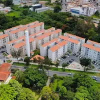 Vista aérea de um conjunto de prédios residenciais de cinco andares com telhado laranja, com estacionamento, playground e duas caixas d’água, ao lado de uma avenida arborizada
