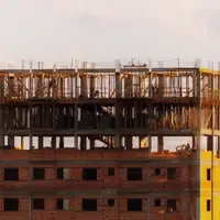 Prédio residencial em obra ao entardecer, com paredes de tijolo vermelho e amarelo à vista, escoramento de madeira no último pavimento e operários de capacete trabalhando no topo