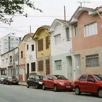 Fileira de casas geminadas de dois andares, cada uma de uma cor, com carros estacionados ao longo de uma rua residencial em ladeira